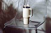 A stylish thermo mug with handle and straw, placed on a transparent acrylic chair, creating a modern and minimalist aesthetic.