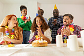 A lively birthday party with a group gathered around a cake. Party hats, colourful wreaths of flowers and smiling faces create a cheerful atmosphere.