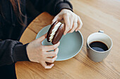 A person enjoys a cake on a plate with a cup of coffee next to it on a wooden table. The scene captures a cosy, enjoyable moment during a break.