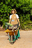 A gardener in a sun hat and apron stands in a lush garden with a wheelbarrow full of vegetables and a tablet that illustrates the connection between agriculture and technology