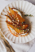 Ein Gourmet-Birnen-Toast mit Birnenscheiben, Käsekrümeln, Honig und Thymian, serviert auf einem stilvollen Keramikteller.