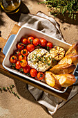 An elegant dish of baked camembert, paired with cherry tomatoes, roasted garlic and toasted baguette slices, garnished with fresh herbs, is a perfect starter.