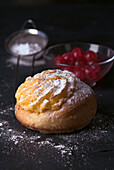 A freshly baked cream roll, generously covered with a layer of velvety cream and dusted with icing sugar, accompanied by a bowl of cherries in the dimly lit setting.