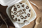 Top view of a beautifully designed bento birthday cake in a white takeaway box, with elegant happy birthday icing, surrounded by chocolate decorations.