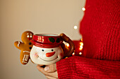 Close-up of a cosy winter scene with a person in a red jumper holding a snowman mug with a gingerbread motif in front of soft, warm lighting