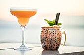 A classic cocktail in a coupe glass and a Moscow Mule in a hammered copper mug sit on a table with a stunning sunset over the sea in the background, creating a perfect tropical holiday backdrop.
