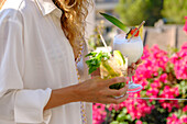 Woman in a white blouse with two summer cocktails, a mojito and a piña colada, in front of a lively garden backdrop. Ideal for concepts of relaxation, summer and leisure.
