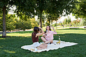Ein fröhliches lesbisches Paar genießt ein sonniges Picknick in einem üppigen Park. Auf einer Decke sitzend, teilen sie Getränke und Lachen und halten den Moment mit einer Kamera fest