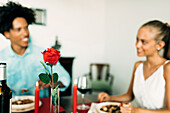 Ein gemütliches, romantisches Abendessen mit einem Paar, das eine Mahlzeit mit einer roten Rose als Mittelpunkt genießt. Warmes Kerzenlicht unterstreicht die intime Atmosphäre und hebt den Wein und das Essen hervor.