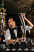 Ein professioneller Barkeeper balanciert gekonnt Cocktail-Shaker und zeigt damit sein Fachwissen und sein Gespür in einer stilvollen Cocktailbar. Martini-Gläser stehen aufgereiht an der Bar, bereit zum Servieren.