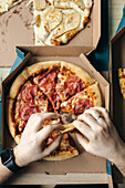 Unrecognisable hand cropped from above, taking various slices of pizza from cardboard boxes, highlighting a casual indoor dining situation, with one person wearing a smartwatch.