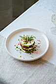 An elegant gourmet salad with fresh red radishes, crunchy cucumber slices, herbs and edible flowers on a white plate next to a glass of water.