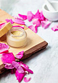 A glass jar with rose face cream and a wooden lid stands on a wooden board, surrounded by bright pink rose petals. In the background is a white mortar, indicating a natural theme.