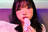 Asian Gen Z girl snacking on candyfloss in an arcade surrounded by glowing neon lights Her playful expression captures the excitement and fun of the urban environment