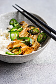 This picture shows, from an oblique angle, a bowl of BBQ tofu slices served on white rice, garnished with fresh green jalapeno slices and accompanied by black chopsticks.