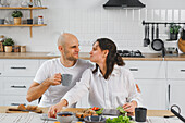 A blind couple have breakfast together in a bright, modern kitchen. They smile at each other, enjoy coffee and fresh sandwiches and create a cheerful and happy morning atmosphere.