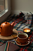 A warm scene with an orange-coloured tea set by the window, a candle, a diary and a blanket. The autumnal ambience is perfect for relaxation and contemplation.
