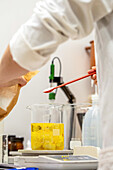 A scientist in a laboratory carefully adds an ingredient to a beaker containing a yellow liquid. The picture shows the precise process of formulating natural cosmetic products.