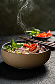 Freshly prepared Vietnamese beef pho noodle soup (Pho Bo) in a ceramic bowl with rising steam over basil, onion, chilli and lime garnishes and chopsticks on the side.