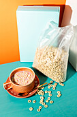 Inviting breakfast scene with a cup filled with milk and cereal, a half-opened cereal box and loose pieces of cereal against a bright blue-green and orange background.