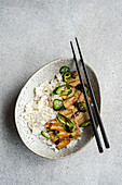 Top view of a dish of grilled tofu slices served on a bed of white rice, garnished with spicy green pepper strips. A pair of black chopsticks on this vegan dish on a ceramic plate against a textured grey background.