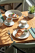 Sunlit breakfast table with butter croissant, latte, hot tea and a bowl of fresh fruit A charming arrangement with natural light and green plants in a cosy café