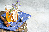 A jar filled with honey, decorated with sprigs of lavender tied together with a white string The scenery includes a honey spoon, old scissors and a blue cloth on a rustic surface