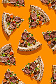 Top view of delicious pizza slices, flavoursomely topped with roast beef, truffle sauce, mozzarella, porcini mushrooms and fresh cherry tomatoes. Each slice is generously garnished with green onions on a bright orange background.