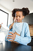 A young girl stands in a modern kitchen and carefully pours water from a jug into a glass. Her casual outfit and the light-coloured kitchen environment reflect a relaxed atmosphere.