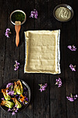 A flat pastry sheet on parchment, surrounded by fresh courgette flowers, purple blossoms and bowls of herb sauce on a dark wooden surface, presenting culinary art.