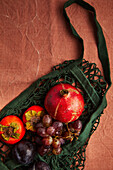 A rustic display of autumn fruits, including pomegranates, grapes, persimmons and plums, arranged in a dark green mesh bag on a textured brown surface.