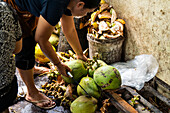 A person skilfully opens coconuts with a knife, surrounded by coconut shells. The setting is a lively, rustic market where traditional preparation methods take centre stage.