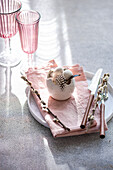An elegant, festive Easter table setting in soft pastel colours. The centrepiece is a decorative egg in the middle, surrounded by flowering willow branches on a pink napkin. Nearby, pink glasses add a touch of glitter.