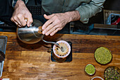 A barista carefully pours water over the coffee grounds in a pourover machine, emphasising the art of preparing speciality coffee. The ingredients nearby add a cosy touch to the wooden worktop.