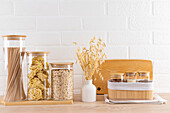 A modern kitchen with a white brick background and transparent storage jars containing pasta, grains and oats, next to a jar of wheat and wooden kitchen utensils.