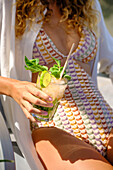 Ein erfrischender Mojito mit Minze und Limette, gehalten von einer Frau in Sommerkleidung. Eine sonnenbeschienene Szene mit farbenfroher Bademode, die das Wesen der Entspannung und der tropischen Stimmung einfängt.