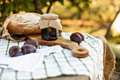 Eine rustikale Einrichtung mit knusprigem Brot, einem Glas hausgemachter Marmelade und reifen Pflaumen auf einem Holzbrett. Perfekte Zutaten für die Zubereitung köstlicher Häppchen.