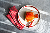Top view of a plate of organic red trout caviar on a toasted slice of bread, accompanied by a red dotted napkin and spoon, presented on a textured grey surface illuminated by natural sunlight.