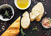 Sliced fresh, crusty baguette with salt, olive oil, olives and basil against a black concrete backdrop