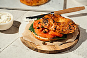Ein frisch gebackener Sesam-Bagel, gefüllt mit Räucherlachs, Kapern und Grünzeug auf einem Holzteller. Leichter Frischkäse wird mit einem Messer aufgestrichen und verleiht ihm eine cremige Textur.