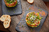From top: two elegant tartare bowls, one with alfalfa shoots, the other simply seasoned, both resting on dark slate slabs. This culinary arrangement is complemented by sliced baguettes on a wooden tabletop, creating a rustic yet sophisticated presentation that is ideal for gourmet interiors.