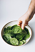 A hand reaches into a bowl of fresh green spinach leaves against a clean white background. The bright green colour is reminiscent of health, freshness and natural goodness.