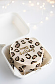 Top view of a stylish bento cake with leopard print and happy birthday decoration in a white takeaway box, surrounded by soft, festive lights.