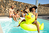 A cheerful girl enjoys a sunny day at the swimming pool and sits on a yellow inflatable duck while her family relaxes nearby. Perfect summer fun, family time and outdoor activity.