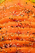 A close-up of thinly sliced smoked salmon, decorated with black and white sesame seeds and herbs, with a bright orange colour and appetising texture.