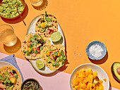 Taco ingredients from above on a bright yellow and pink background. The picture shows tacos topped with salmon, avocado, coriander and mango. They are served with a bowl of guacamole, pumpkin seeds, salt and lime slices.