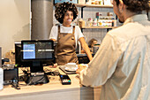A cheerful café employee with vitiligo in an apron serves a customer at the counter The pleasant ambience and professional service emphasise the inviting environment of the café