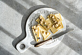 Top view of a sliced white chocolate bar with nuts, accompanied by a rustic knife on a decorative ceramic plate, beautifully illuminated by natural light against a concrete background