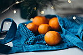 Fresh mandarins in a bright blue string bag against a softly lit festive backdrop create a festive and cosy atmosphere. The blue bag provides a colour accent that emphasises the orange tones of the mandarins.