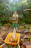 A gardener stands smiling in a vegetable garden, holding a basket in his hand. The sunlight falls through the trees and highlights the tools in a wheelbarrow, symbolising sustainable agriculture
